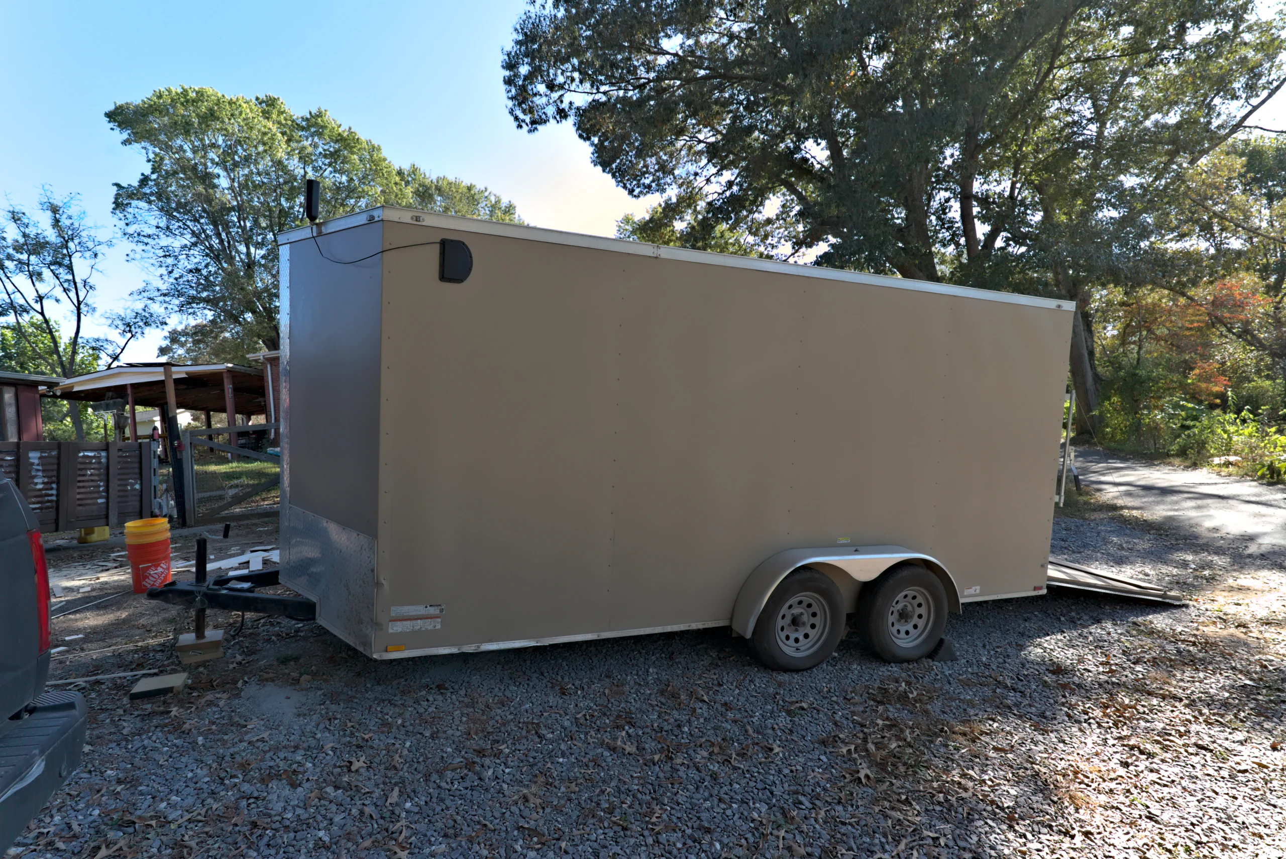 Side view of the tan enclosed V-nose Biltout prototype trailer parked on gravel, with its ramp down at the rear