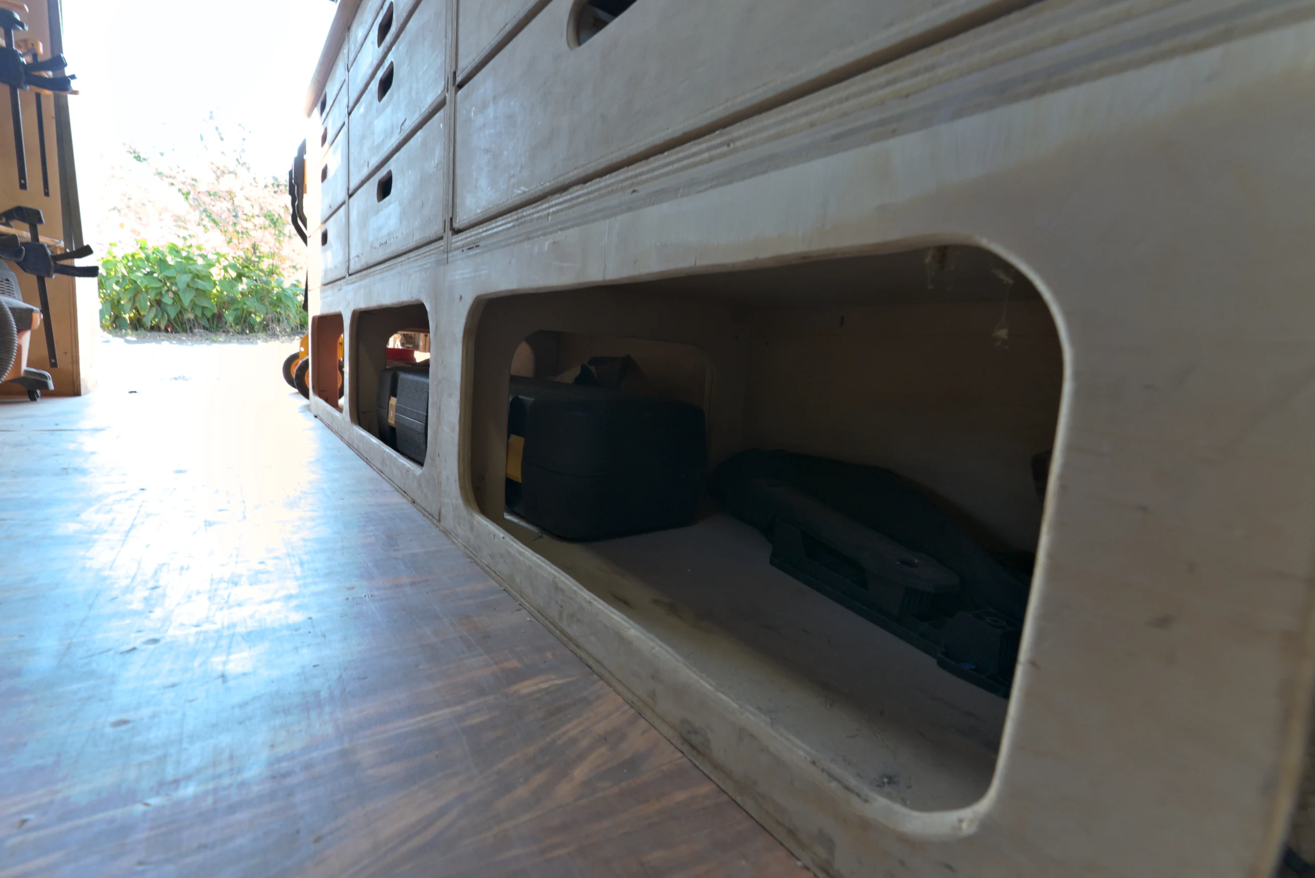 Low plywood storage bays with oval openings holding tool cases, seen from floor level along the aisle of the Biltout prototype